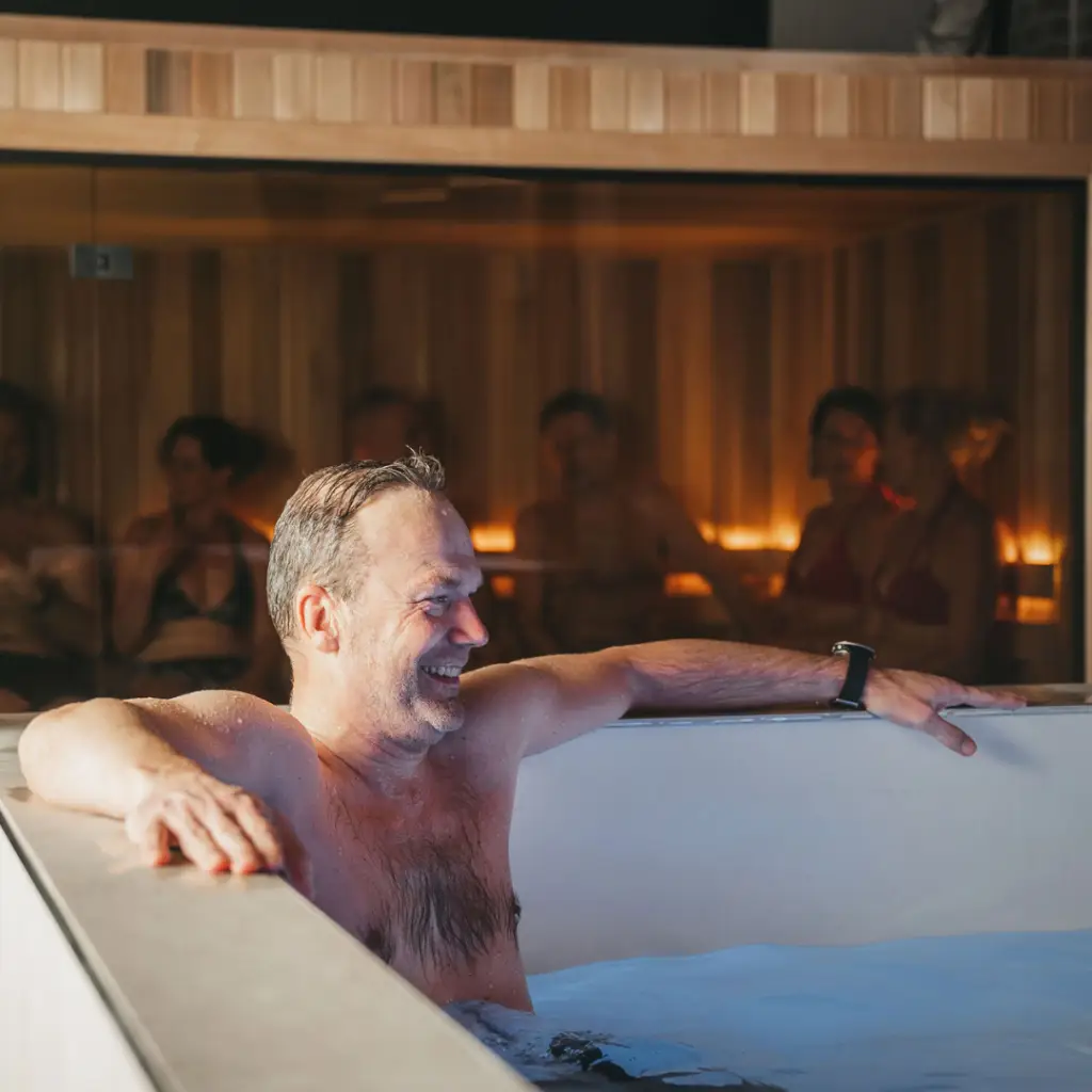 Bij COMMIT Utrecht geniet een man van een ontspannend bad in de hot tub; hij glimlacht tevreden terwijl zijn armen comfortabel op de rand rusten. Zachte verlichting en uitnodigende houten wanden zorgen voor de juiste sfeer terwijl gasten samen tot rust komen in de sauna en genieten van de warmte en het buitengewone comfort van onze eersteklas healthclub-ervaring.