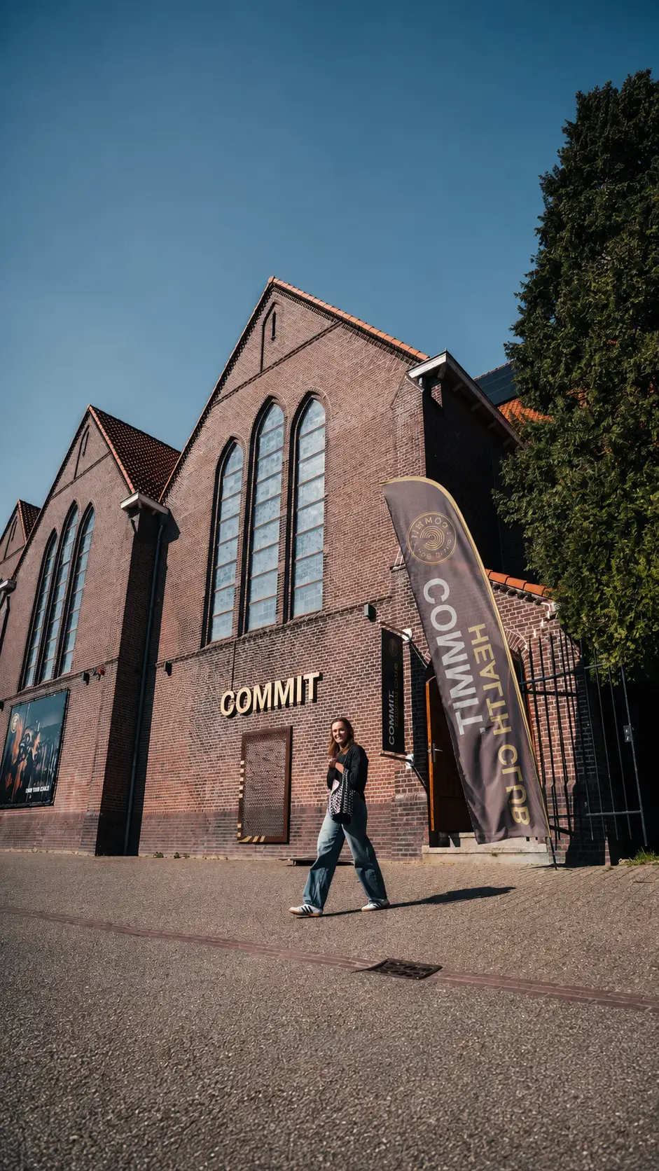 Een gast wandelt langs ons elegante, bakstenen clubhuis, dat opvalt door de boogramen en het opvallende COMMIT-bord. Bij de ingang prijkt een opvallende banner met de tekst ‘HEALTH CLUB COMMIT’, waarop onze tarieven voor de premium sportschool in Utrecht worden uitgelicht. Weelderige bomen omlijsten het gebouw en zorgen voor een gastvrije sfeer voor alle bezoekers.
