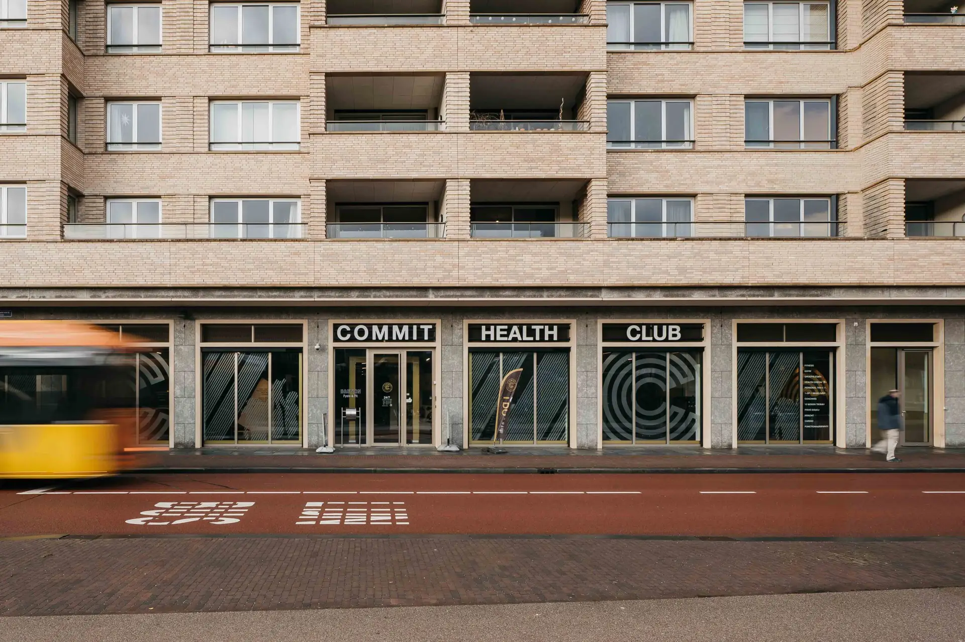 Aan de voet van een stijlvol beige appartementencomplex staat Commit Health Club klaar om je te verwelkomen met faciliteiten van topkwaliteit en persoonlijke begeleiding. Buiten glijdt een gele bus soepel door de levendige straat, terwijl een voorbijganger langs onze uitnodigende ingang slentert – een momentopname van het bruisende stadsleven, op slechts een steenworp afstand van je volgende training.