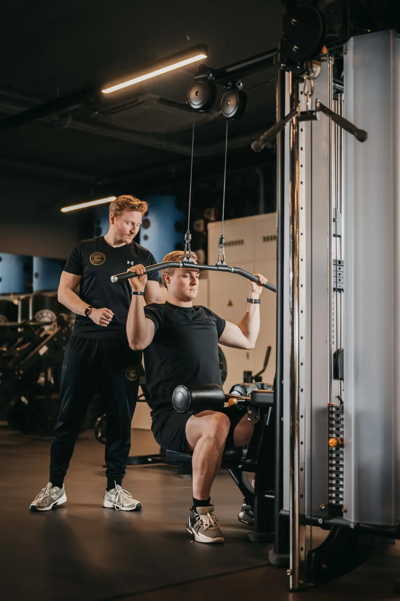 Bij COMMIT Health Club Utrecht voert een lid onder de aandachtige begeleiding van zijn personal trainer een gerichte lat pulldown uit. Beiden zijn gekleed in strakke zwarte sportkleding en belichamen daarmee ons streven naar fitness van topkwaliteit in een gastvrije, ondersteunende omgeving. Elke sessie wordt vakkundig begeleid, zodat je je doelen met vertrouwen en zorg kunt bereiken.
