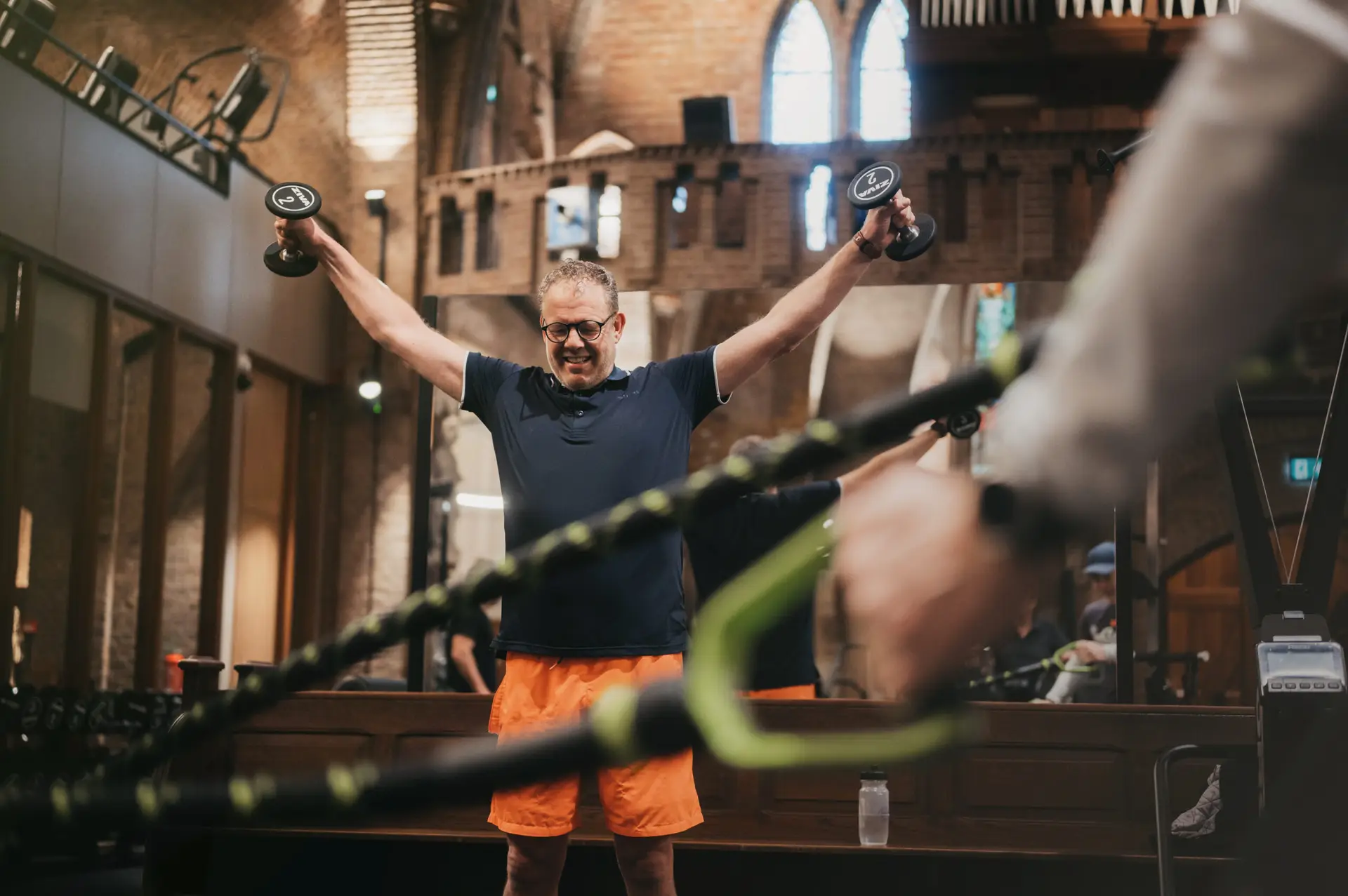 Een man in een uitnodigende sportschoolomgeving tilt halters op met beide armen omhoog; hij draagt een marineblauw shirt, een oranje korte broek en een bril. De ruimte valt op door de hoogwaardige bakstenen muren en elegante houten accenten – kenmerken van de persoonlijke, professionele sfeer bij COMMIT FIT in Utrecht. Op de voorgrond traint een andere bezoeker zelfstandig, maar wordt daarbij ondersteund door deskundige coaching op ultramoderne apparatuur.