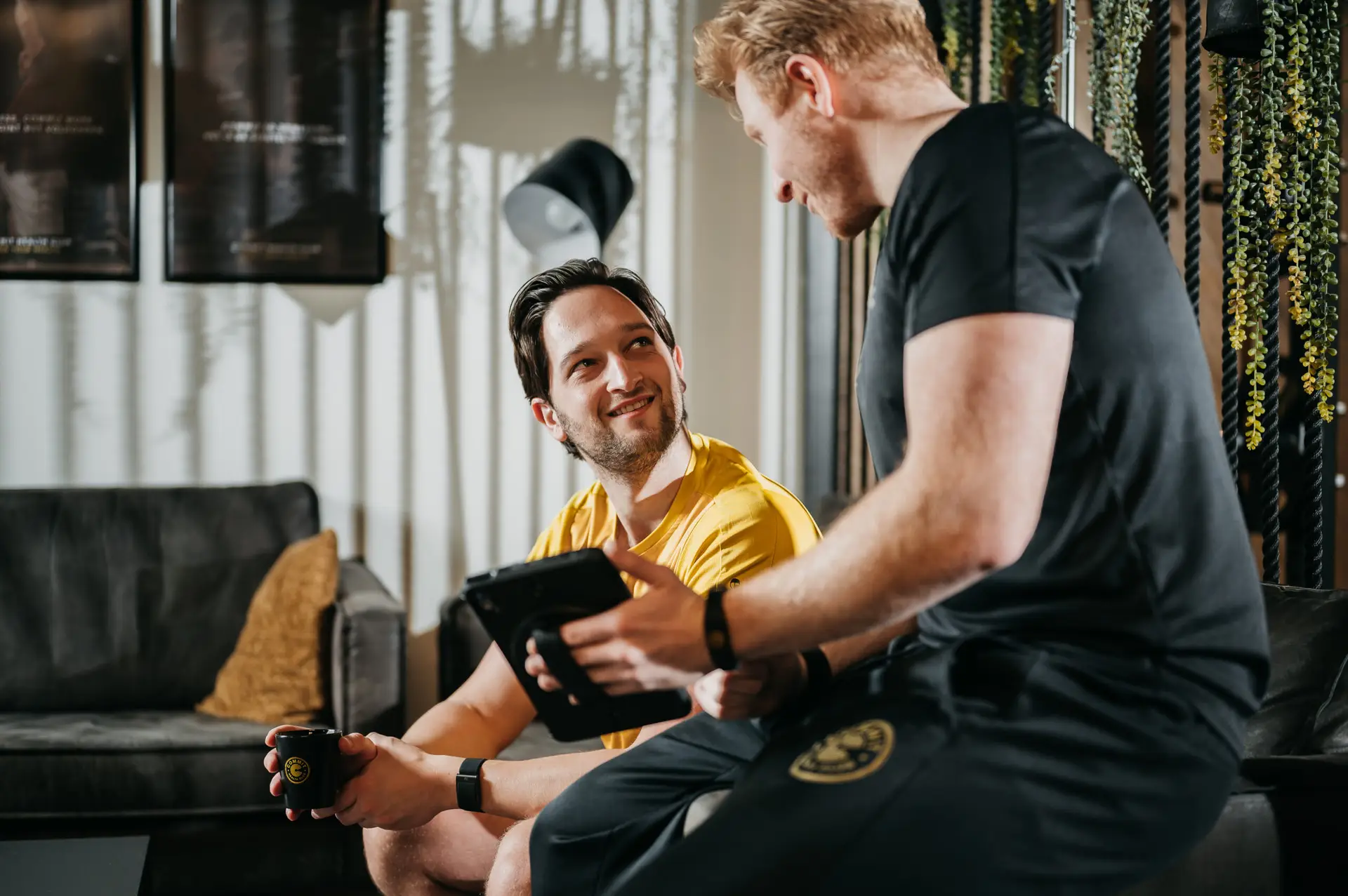 Twee mannen zitten samen binnen; de ene draagt een geel shirt en houdt een mok vast, de andere is in het zwart gekleed en heeft een tablet bij zich. Beiden glimlachen oprecht en zijn volledig in gesprek. Achter hen zorgen weelderige planten en een comfortabele bank voor een uitnodigende sfeer – die de hoogwaardige, persoonlijke en professionele omgeving van COMMIT Health Club weerspiegelt. Hier weerspiegelt elk detail onze toewijding aan fitnessbegeleiding op maat, afgestemd op uw unieke doelen.