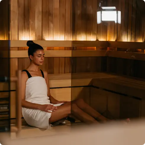 Een vrouw, gehuld in een witte handdoek, zit rustig in een warm verlichte, houten sauna bij COMMIT Utrecht. Met haar ogen dicht geniet ze van de rustgevende, exclusieve sfeer – een kenmerk van onze healthclub in Utrecht, waar persoonlijke aandacht en professionele begeleiding de basis vormen voor elke wellnesservaring.