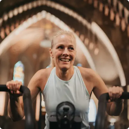 Een vrouw in een mouwloos topje geniet van haar training op moderne fitnessapparatuur bij COMMIT Health Club Utrecht. Het elegante, gewelfde interieur met prachtige glas-in-loodramen zorgt voor een inspirerende en uitnodigende sfeer, die de unieke combinatie van eersteklas fitness en deskundige begeleiding weerspiegelt die onze club kenmerkt.