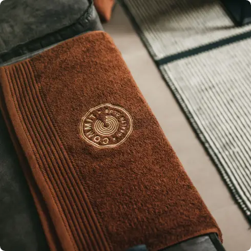 Een luxe bruine handdoek, elegant opgevouwen en versierd met het kenmerkende ronde logo en de huisstijl van COMMIT Health Club Utrecht, ligt op een strak donker oppervlak naast een zacht beige-zwart gestreept vloerkleed, wat onze toewijding aan comfort en kwaliteit weerspiegelt.