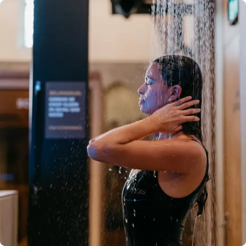 Een vrouw met nat haar geniet van een verfrissende douche bij COMMIT Health Club Utrecht, terwijl ze haar handen zachtjes op haar nek laat rusten onder het stromende water. De rustige omgeving weerspiegelt onze kenmerkende combinatie van eersteklas faciliteiten en een gastvrije sfeer, en biedt een serene ervaring bij de toonaangevende luxe sportschool van Utrecht.