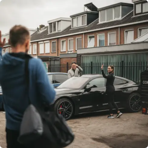 Een man met een plunjezak zwaait hartelijk naar een glimlachende man en vrouw die naast een strakke zwarte auto staan voor een rij charmante huizen, klaar voor hun bezoek aan een luxe fitnessclub in Utrecht, waar gepersonaliseerde en professionele fitnessbegeleiding op hen wacht.