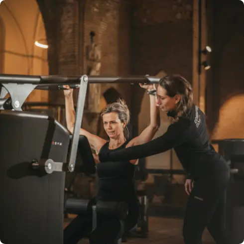 Bij COMMIT Health Club Utrecht maakt een lid vol zelfvertrouwen gebruik van de shoulder press-machine, terwijl een van onze deskundige trainers naast haar staat om haar in onze eersteklas en gastvrije sportschool aandachtig te begeleiden en aan te moedigen.