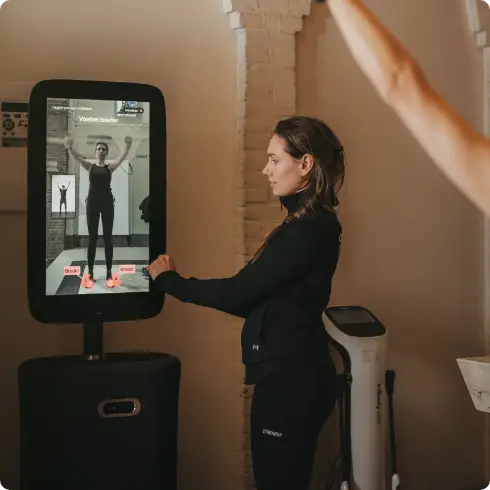 In de uitnodigende sfeer van COMMIT Health Club Utrecht geniet een vrouw van een op maat gemaakte trainingssessie voor een geavanceerde fitnessspiegel, die onze toewijding aan wellness van topkwaliteit weerspiegelt. Aan de rechterrand is de opgeheven arm van een ander lid te zien, wat verwijst naar een gedeelde ervaring binnen onze levendige gemeenschap.