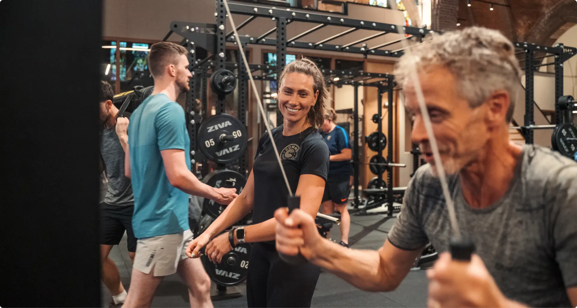Leden genieten van een dynamische trainingssessie in onze eersteklas sportschool; een vrouw glimlacht zelfverzekerd terwijl ze de kabelmachine gebruikt, terwijl een man op de voorgrond bezig is met een ander apparaat. Op de achtergrond zijn anderen bezig met hun fitnessroutines, allemaal in onze gastvrije en professionele sportschoolomgeving.