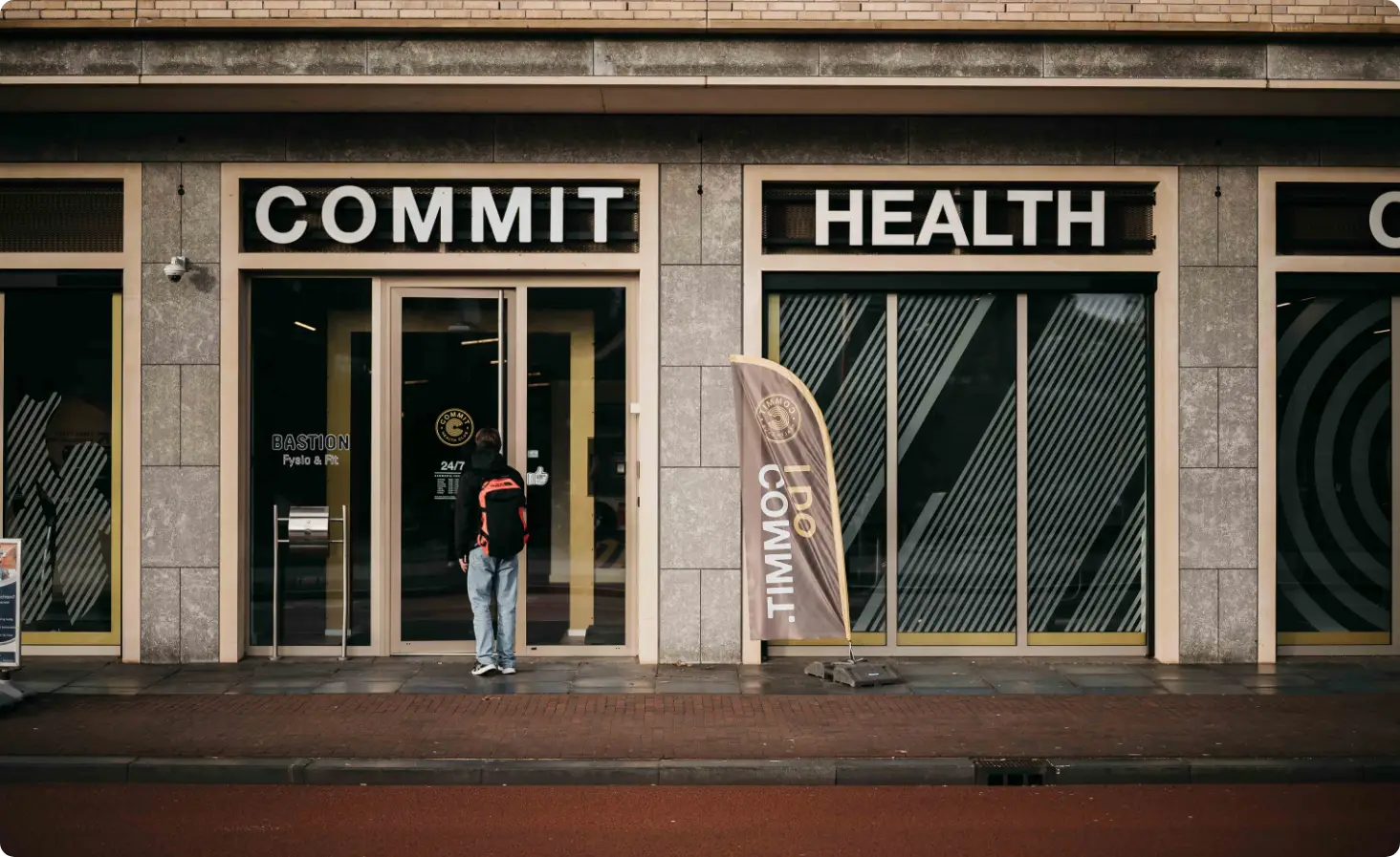 Bij de ingang van een strak COMMIT HEALTH-gebouw staat een gastvrije medewerker, omringd door indrukwekkende glazen ramen en onze kenmerkende Commit 100%-vlag – een symbool van onze toewijding aan uw welzijn. De uitnodigende wandelroute en het uitzicht op de straat leiden u naar een van de eersteklas locaties van COMMIT Health Club in Utrecht, zoals Leidsche Rijn of Rivierenwijk, waar persoonlijke aandacht en uitmuntendheid op u wachten.