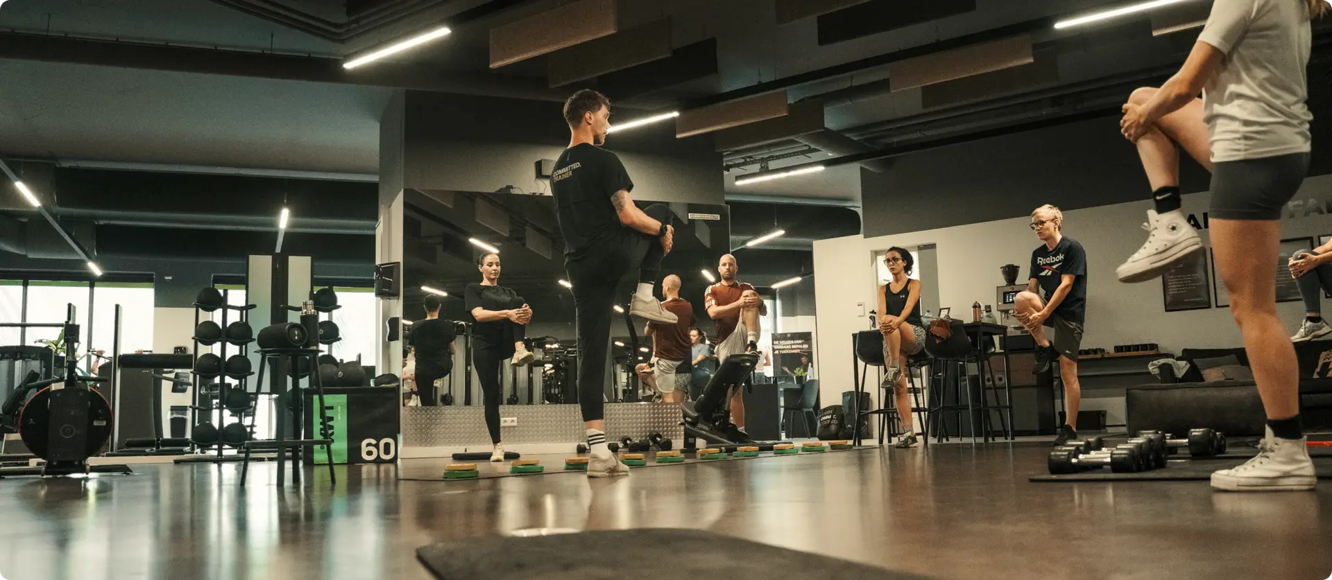 In de uitnodigende, stijlvolle omgeving van een moderne sportschool geniet een dynamische groep, gekleed in hoogwaardige sportkleding, van een motiverende groepsles. Een inspirerende trainer leidt de les met vakkennis, terwijl anderen actief meedoen of aandachtig toekijken. Hieruit blijkt het gevoel van verbondenheid en professionaliteit dat kenmerkend is voor de levendige gemeenschap bij COMMIT Health Club-vestigingen zoals Rivierenwijk en Leidsche Rijn.